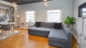 A modern living room with a grey sectional sofa, a dining area with a round table and chairs, and large windows providing natural light. - Bateman, modern City centre apartment sleeps 6 (York)