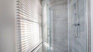 A shower with glass doors and a tiled wall, a window with blinds, and a wooden floor. - Bateman, modern City centre apartment sleeps 6 (York)