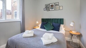 A neatly made bed with a grey and white checkered bedspread, two pillows, and two folded towels on the side. There are two bedside lamps on wooden nightstands. The room has a window with curtains, a wall with framed pictures, and a green upholstered headboard.
