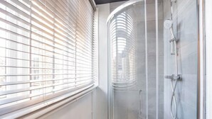 A modern bathroom with a glass shower enclosure, a window with blinds, and a visible showerhead.