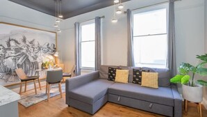 A modern living room with a grey sectional sofa, a dining table with chairs, and a large window with curtains.
