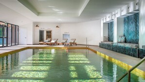 An indoor swimming pool with clear water, lounge chairs, and a seating area with patterned cushions.
