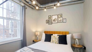 Habitación de hotel con una cama grande, dos mesitas de noche con lámparas, una ventana con cortinas y cuadros en la pared.