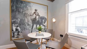 Un comedor con una mesa redonda, cuatro sillas, un cuadro blanco y negro enmarcado, una planta en maceta y una ventana con luz natural.
