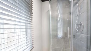 Un baño moderno con ducha de vidrio, bañera blanca y ventana con persianas.
