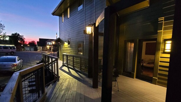 Une terrasse en bois avec une rambarde, un bâtiment doté d’une grande fenêtre et une rue où est garée une voiture.