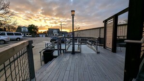 Une terrasse en bois avec une rambarde en métal, un lampadaire et, en arrière-plan, une vue sur des voitures et des maisons garées.
