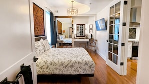 Une chambre à coucher comprenant un lit, un bureau, une chaise, un téléviseur et une porte donnant sur la cuisine.