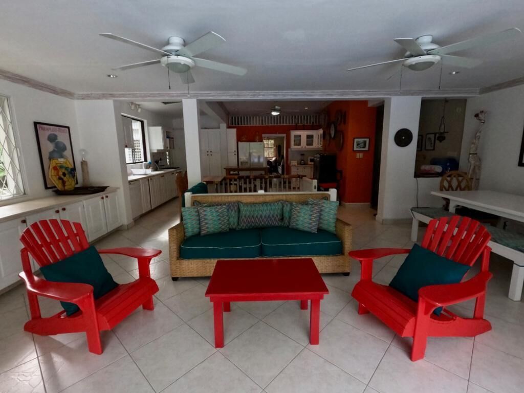Un salon avec des fauteuils rouges et une table rouge, un canapé avec des coussins à motifs, et une cuisine en arrière-plan.
