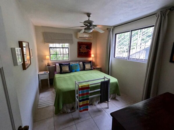 A bedroom with a bed, a nightstand, a dresser, a window with curtains, a ceiling fan, and a view of greenery outside.