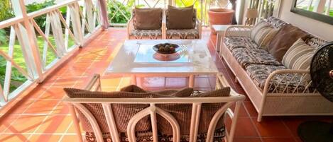 A covered outdoor seating area with wicker furniture, a table with a bowl of fruit, and a view of greenery.