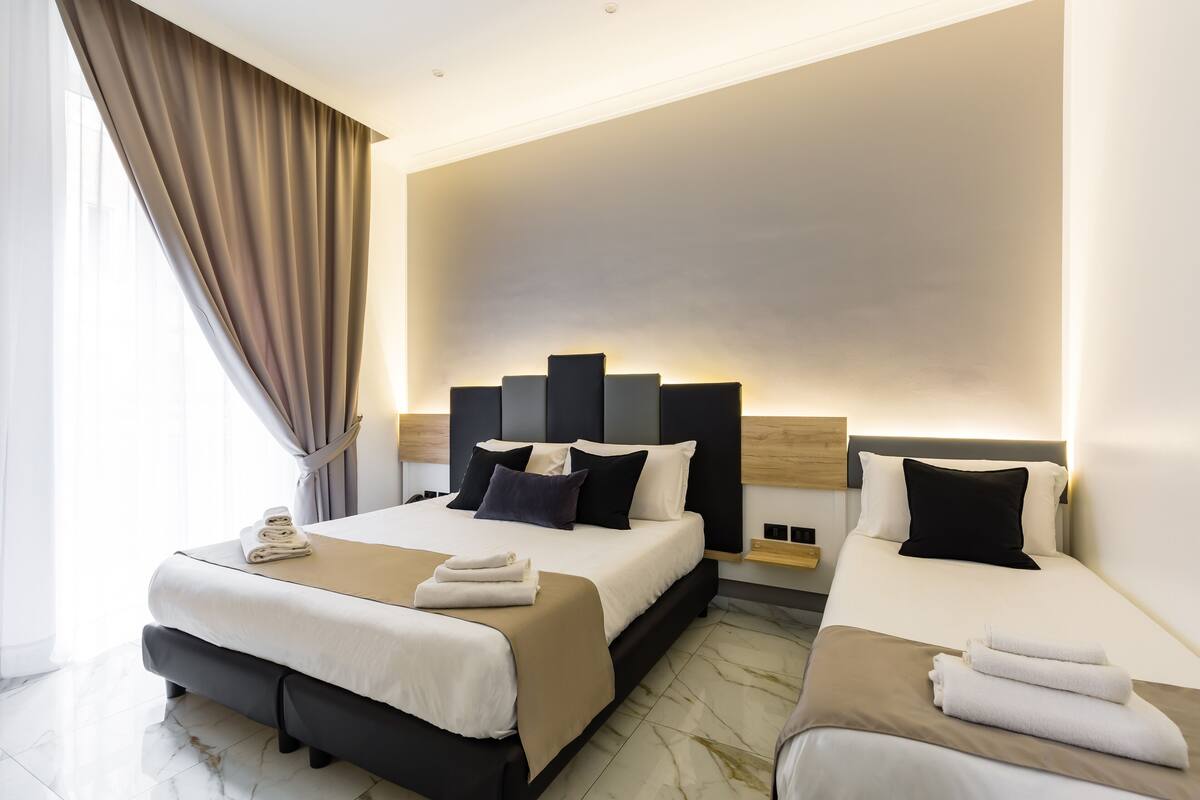 Une chambre d’hôtel avec deux lits, une grande fenêtre avec des rideaux, une tête de lit dotée d’un éclairage intégré et une table de chevet avec une lampe.