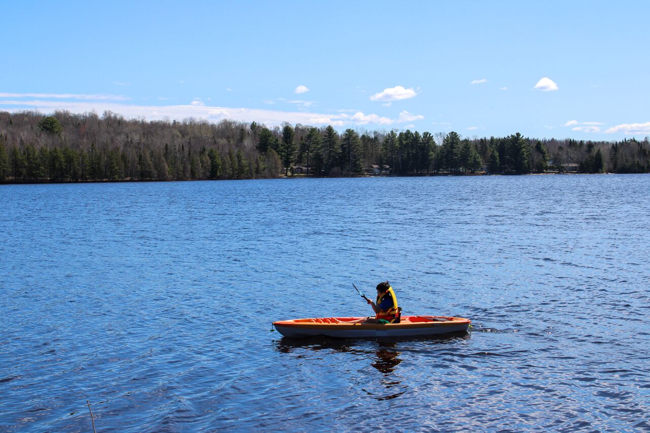 Un kayakiste dans un petit kayak orange sur un lac calme, bordé d’une rive boisée.