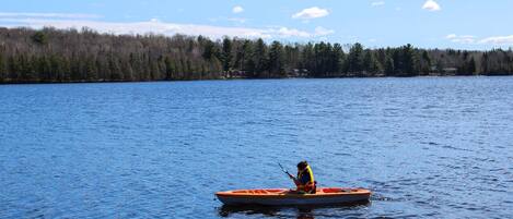 Un kayakiste dans un petit kayak orange sur un lac calme, bordé d’une rive boisée.