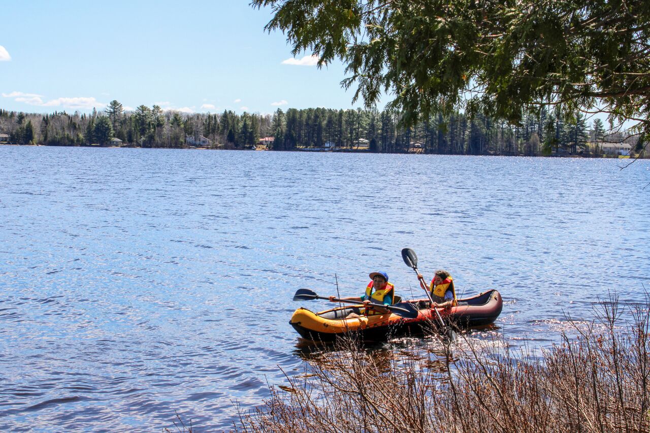 Two people kayaking on a calm lake with trees and a clear sky in the background.