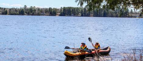Two people kayaking on a calm lake with trees and a clear sky in the background.