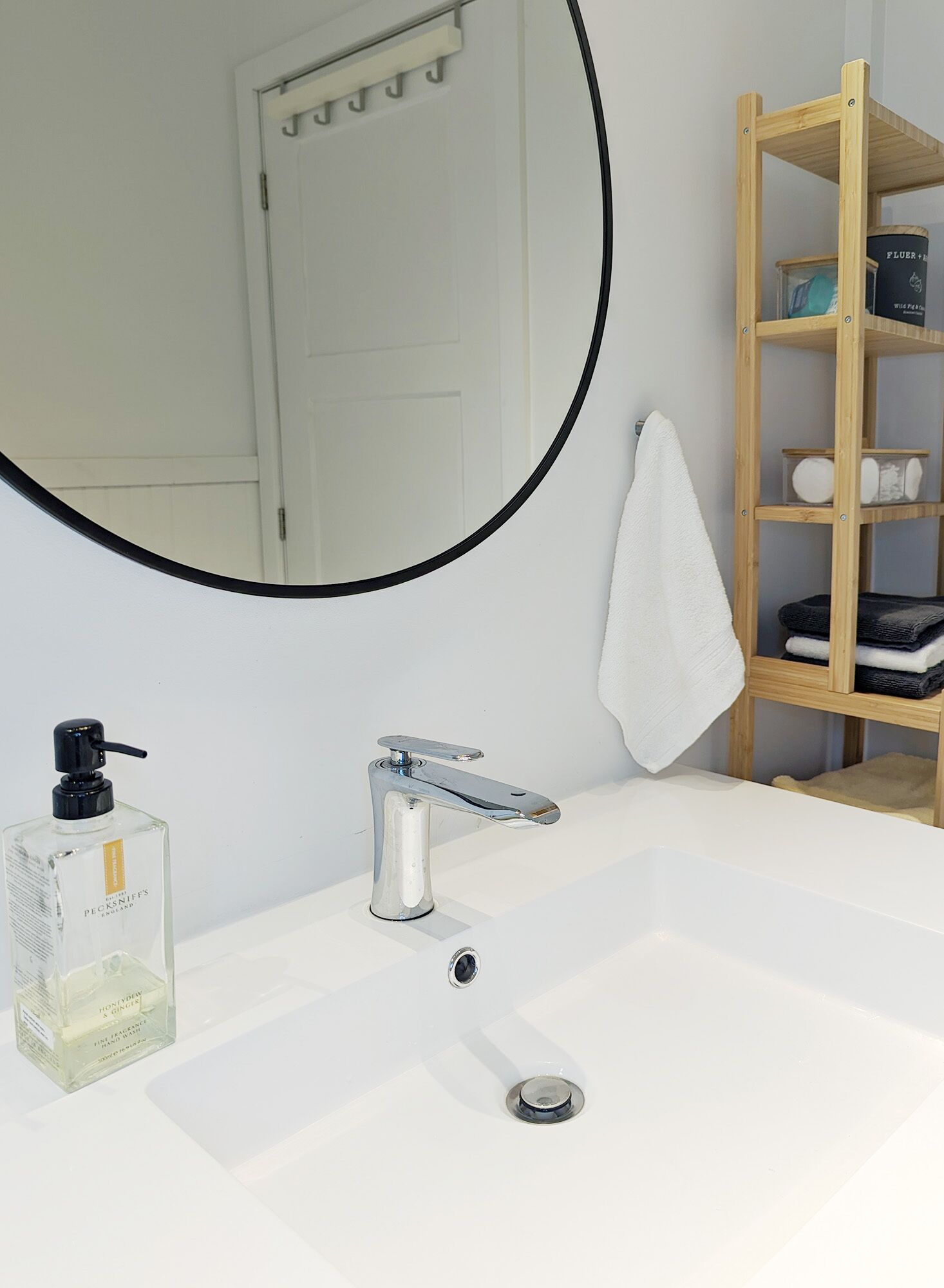Ein Badezimmer mit einem weißen Waschbecken, einem Spiegel mit schwarzem Rahmen, einer Holzablage mit Handtüchern und einer Handseifenflasche.