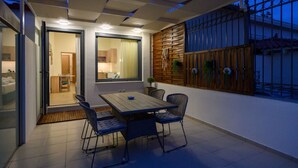 An outdoor patio with a table and chairs, a sliding glass door leading to a kitchen, and a wooden fence with plants.