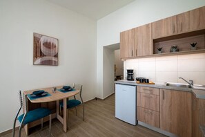 A compact kitchen with wooden cabinets, a small dining table with blue chairs, a coffee maker, and a dishwasher. - Apartment with Sea View (Kalamata)