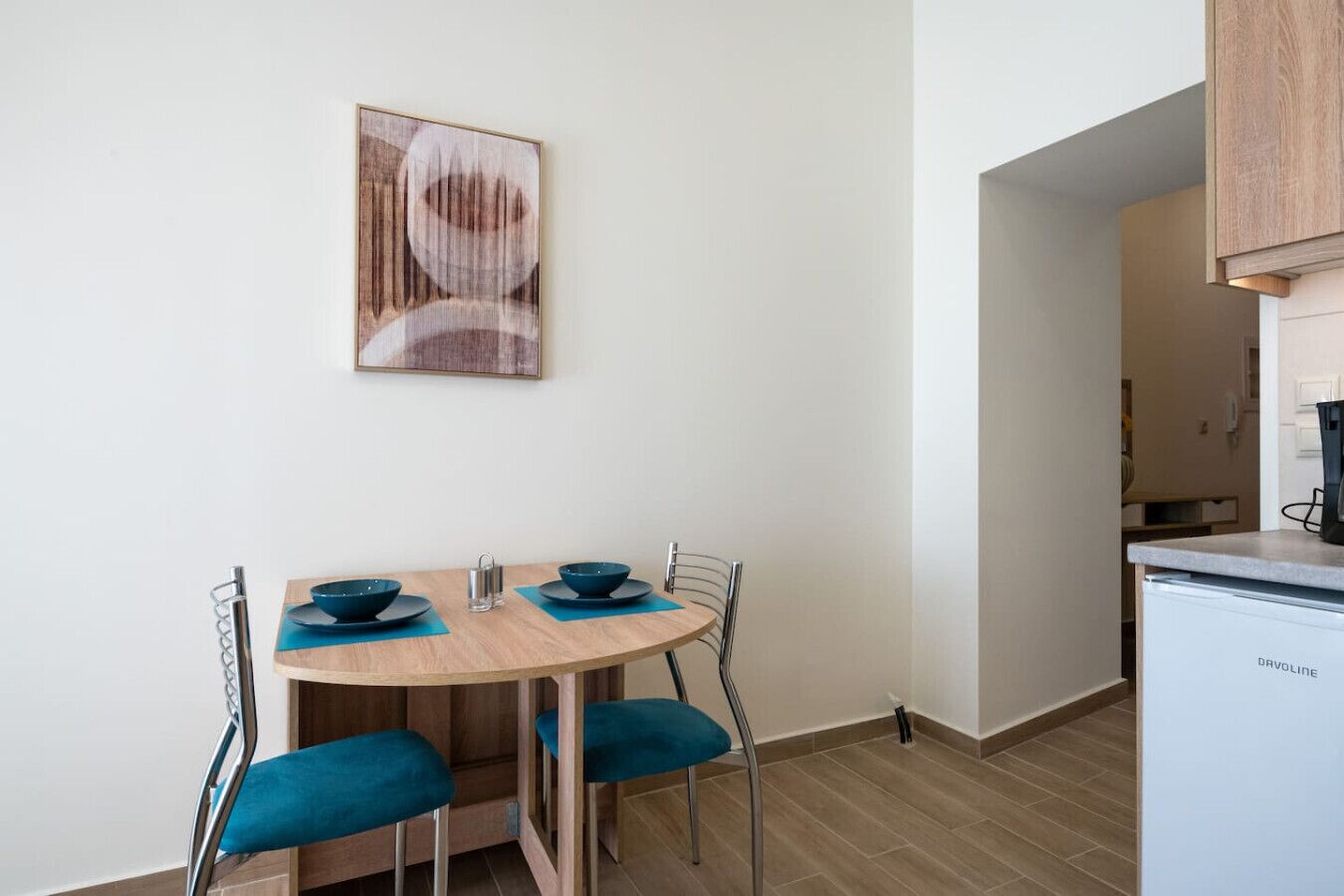 A compact kitchen with a white refrigerator, a wooden dining table with blue chairs, and a framed abstract painting on the wall.