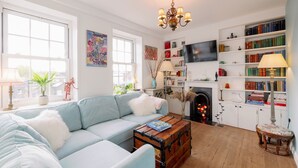 Een gezellige woonkamer met een lichtblauwe bank, een houten kist en een open haard. Er staan boekenkasten, een televisie en een kroonluchter.