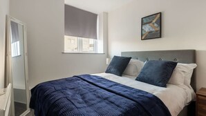 Un dormitorio con una cama, una ventana con persiana, un cuadro en la pared y una puerta a la izquierda.