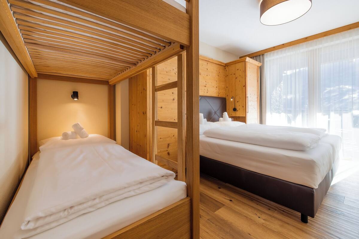 Habitación de hotel con techo de madera, cama con ropa blanca, mesita de noche y un amplio ventanal con cortinas translúcidas.