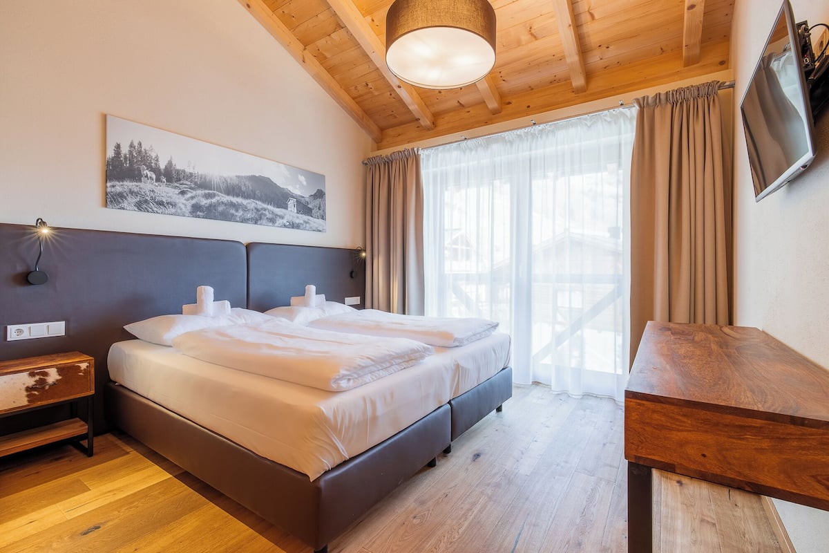 A bedroom with a bed, a wooden desk, a TV, and a window with curtains.