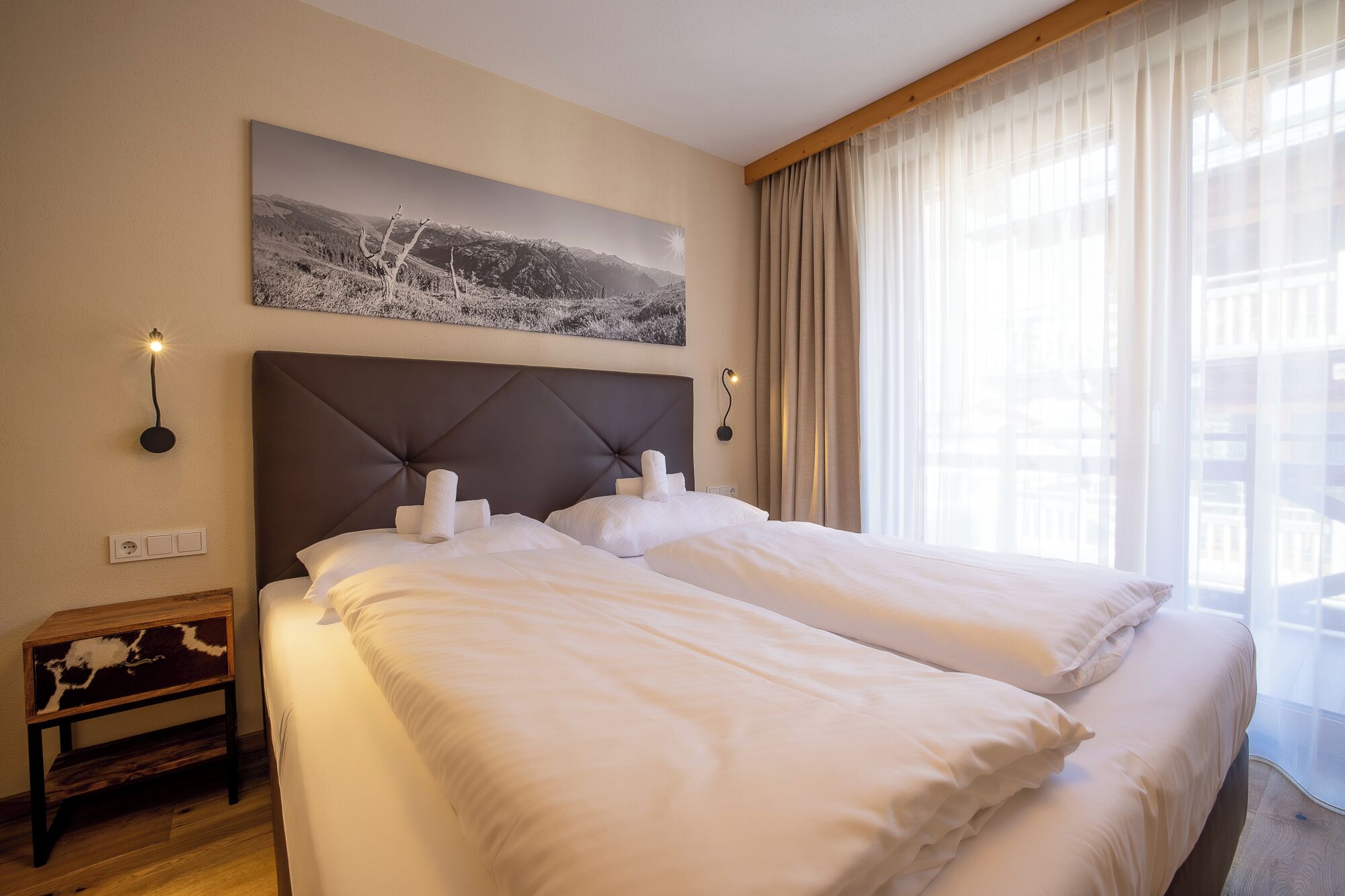 Habitación de hotel con una cama grande, cabecero, mesita de noche, lámpara de pared y ventana con cortinas.