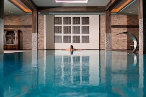 A large indoor swimming pool with a person swimming, surrounded by stone walls and wooden beams.