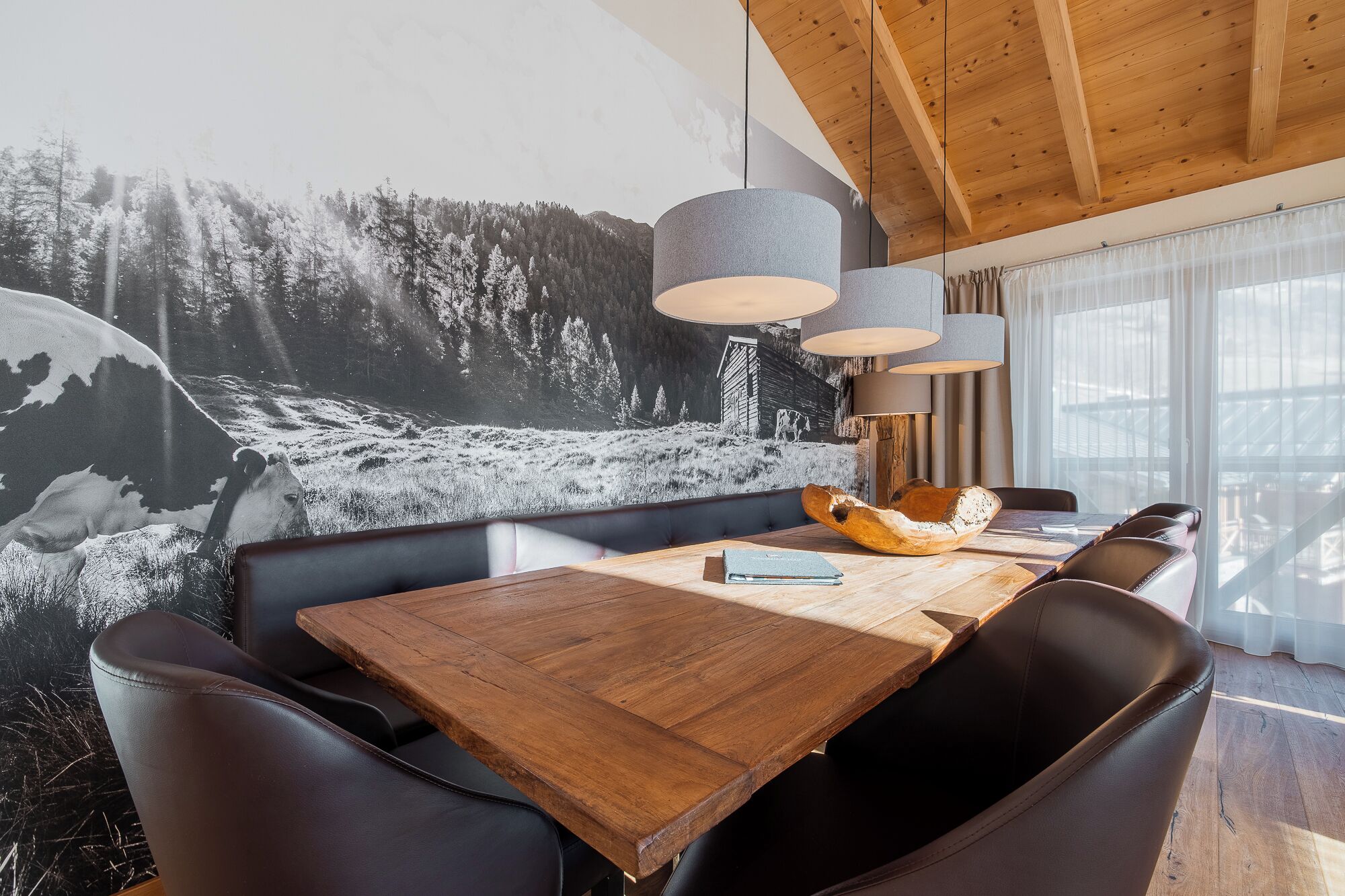 Un coin repas avec une table en bois, des chaises en cuir et une fresque murale représentant un paysage de montagne.