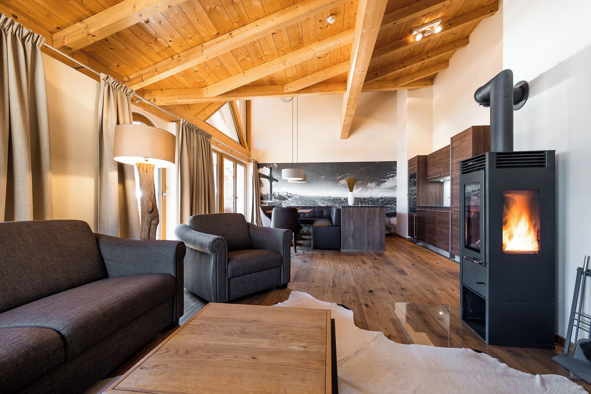 A cozy living room with a fireplace, wooden ceiling beams, and a large window.