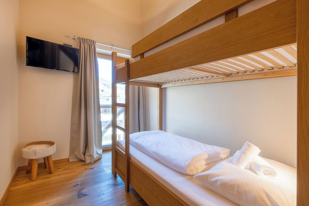 Habitación con literas, un televisor montado en la pared, un taburete de madera y una ventana con cortinas.