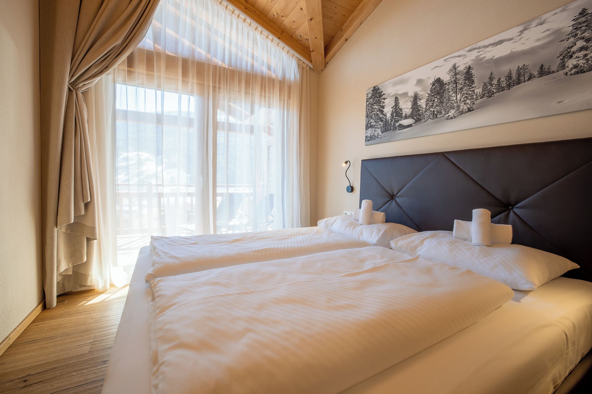 Hotelkamer met twee bedden, een groot raam met dunne gordijnen, een schilderij van een besneeerd landschap aan de muur en een houten plafond.