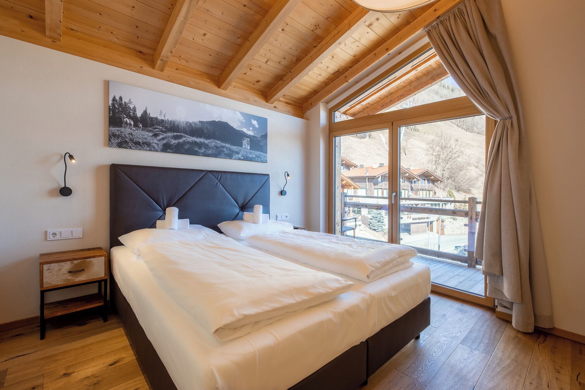 Une chambre avec un grand lit, des poutres en bois au plafond, un tableau au mur et une vue sur une montagne et des bâtiments à l’extérieur.