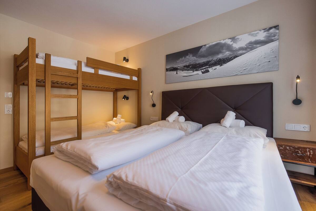 Una habitación con dos camas, una litera de madera y otra con cabecera. Hay un cuadro en la pared y una lámpara en la mesita de noche.