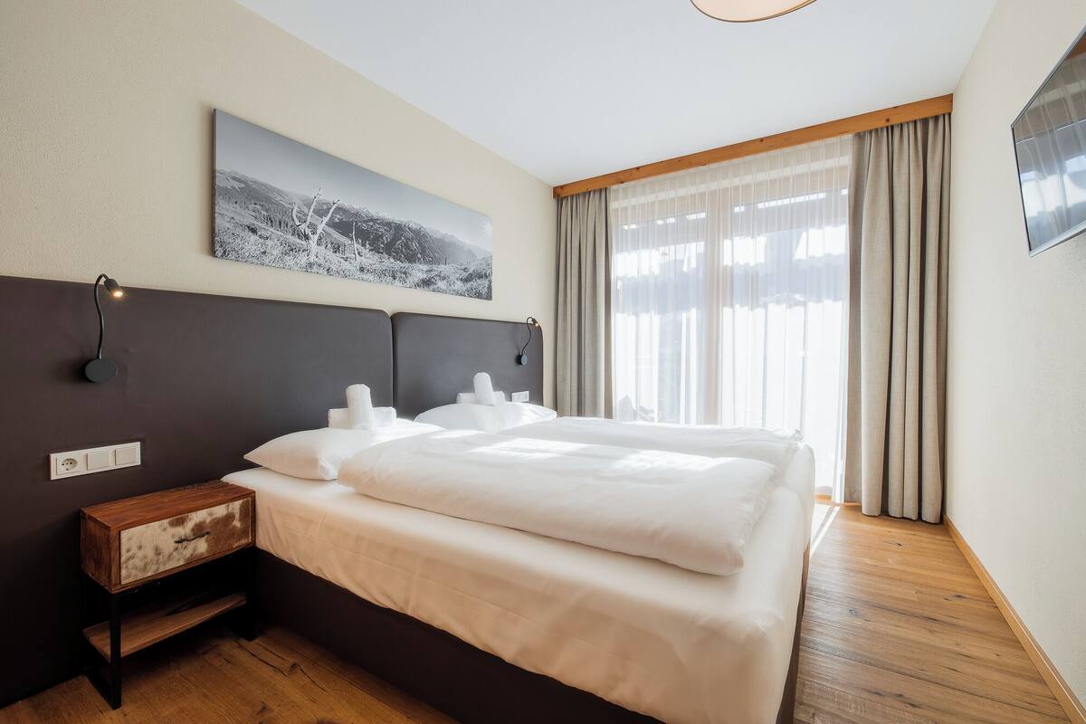Una habitación de hotel con una cama grande, una mesita de noche de madera, una lámpara de pared y un ventanal con cortinas.
