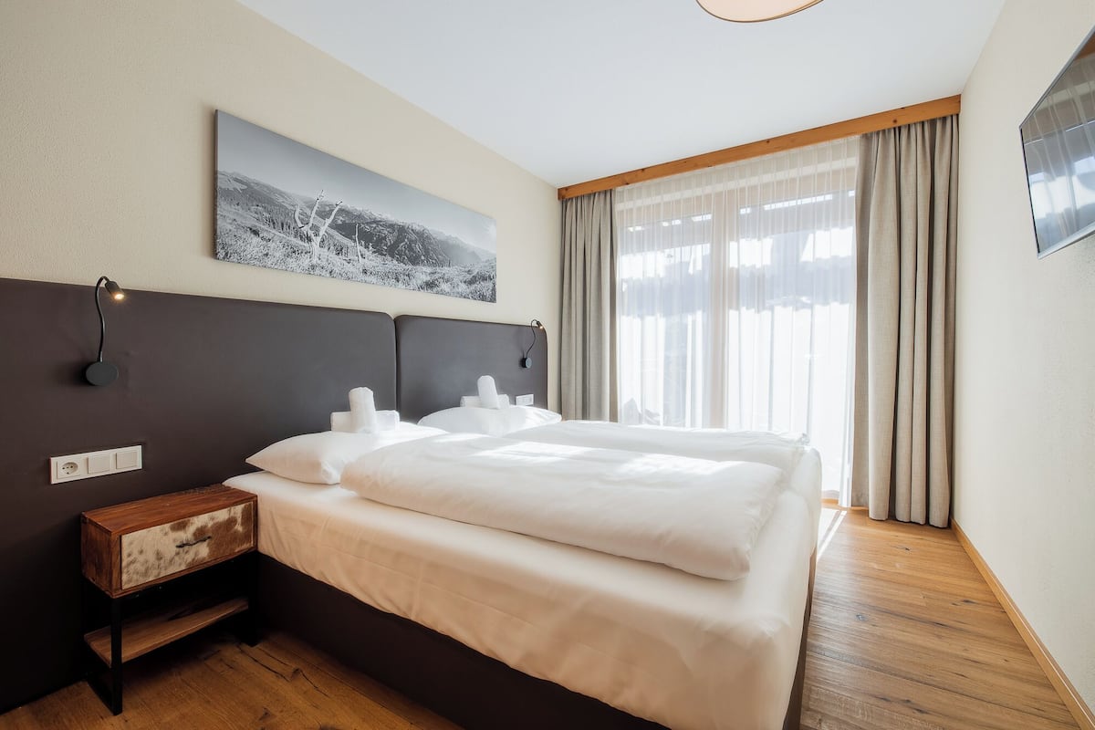 A hotel room with a large bed, a wooden nightstand, a wall-mounted lamp, and a window with curtains.