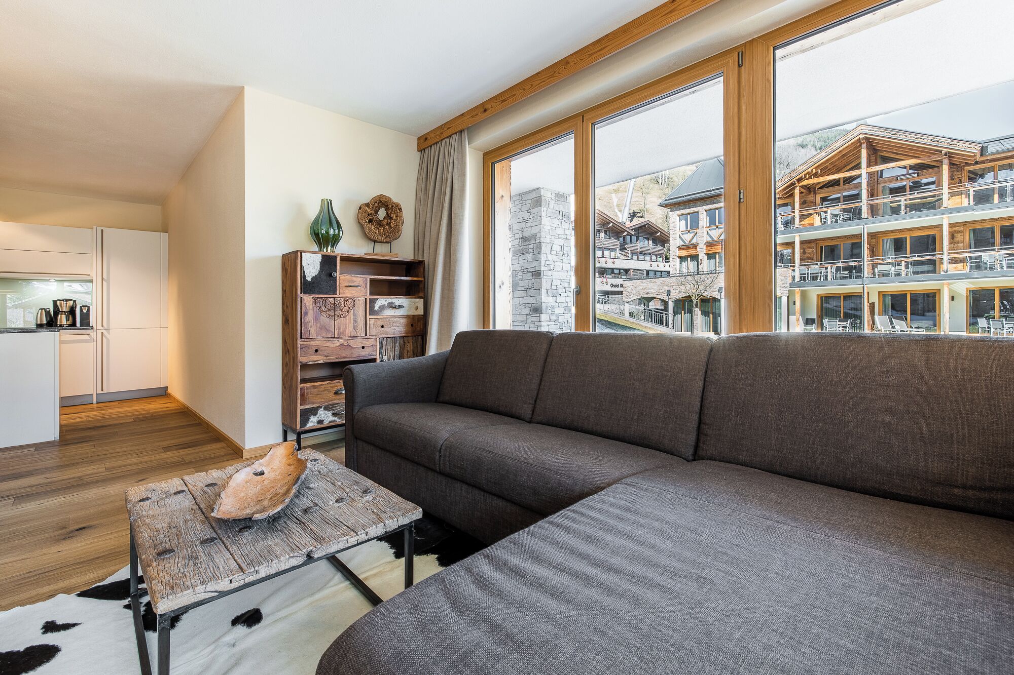 Een moderne woonkamer met een grote grijze bank, een houten salontafel en uitzicht op een meerlaags gebouw met balkons.