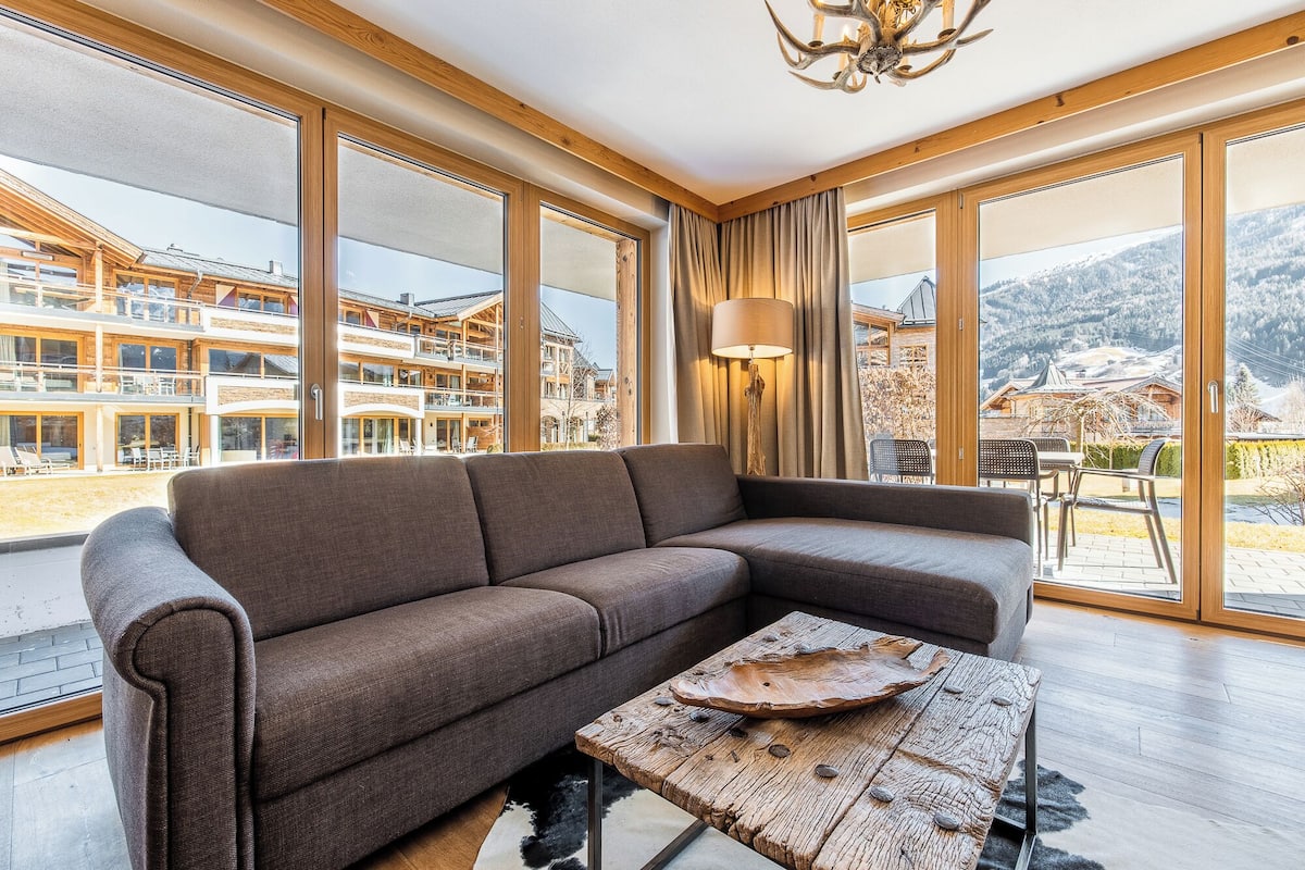 A modern living room with a large sectional sofa, a wooden coffee table, and large windows offering a view of a mountain resort.