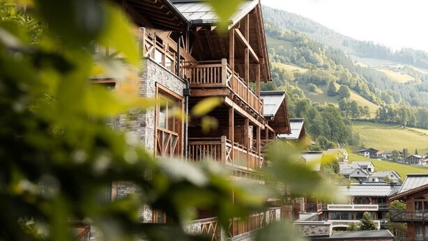 Ein hölzernes Gebäude mit Balkon, eingebettet in eine ländliche Landschaft mit Hügeln und Bäumen.