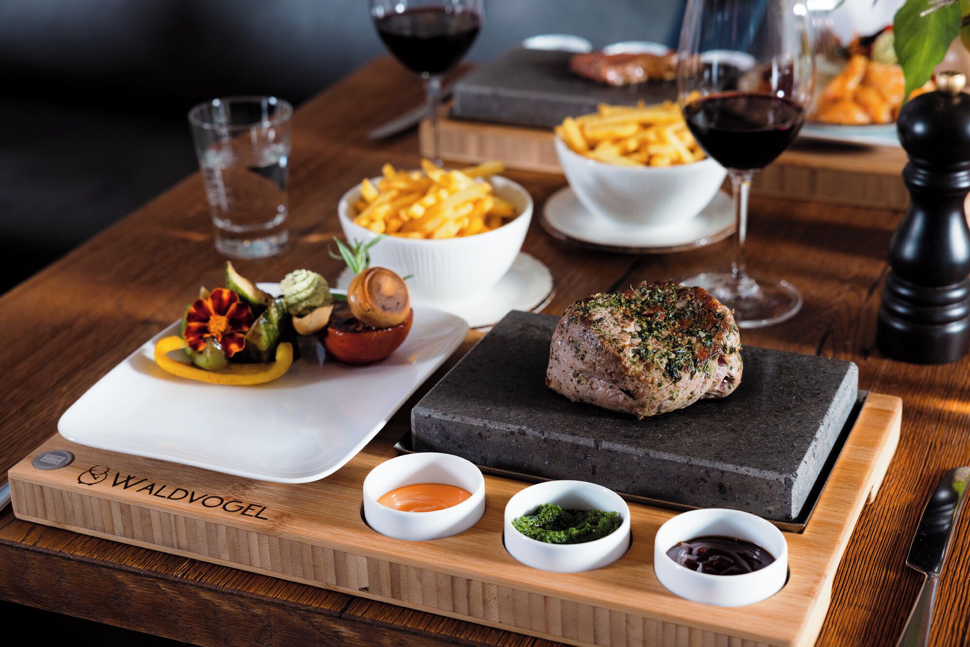 A wooden serving board with a steak, vegetables, and sauces.