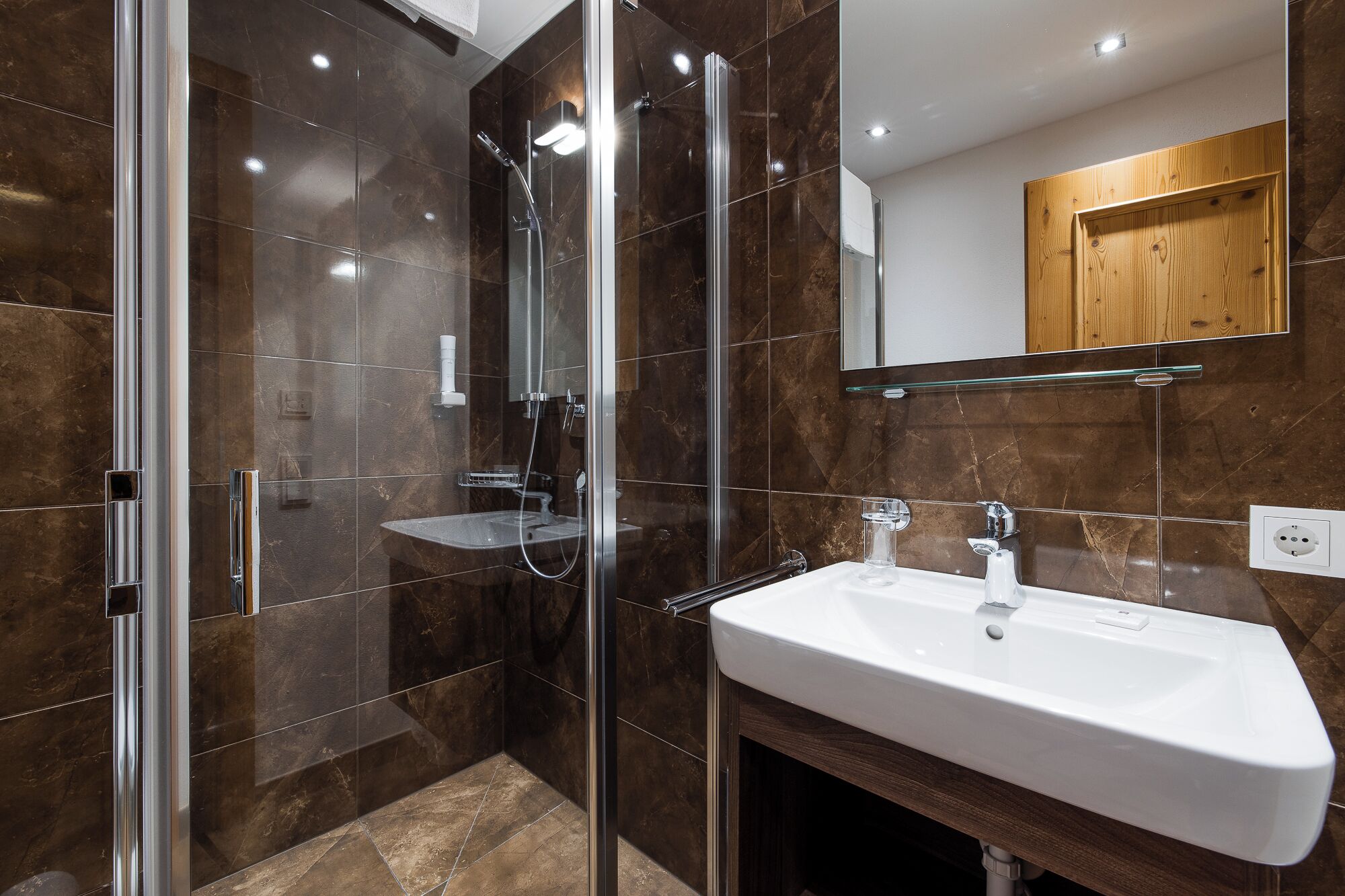 Un baño moderno con ducha de vidrio, lavamanos blanco y mueble de madera.
