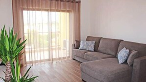 A living room with a brown sofa, two patterned cushions, a large potted plant, and a sliding glass door with sheer curtains. - Apartment Amelo, fantastic clean apartment near marina (Albufeira)