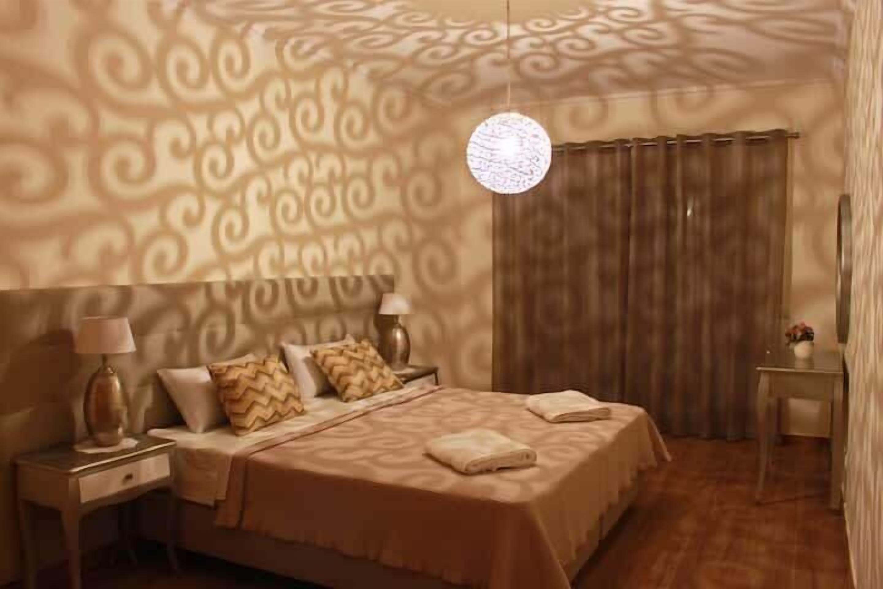 Une chambre d’hôtel avec un papier peint à motifs, un lit recouvert d’une courtepointe beige, deux lampes de chevet, une table de chevet, un bureau et un luminaire suspendu.