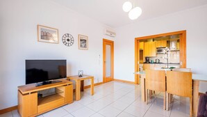 Un salon moderne doté d’un téléviseur à écran plat, d’un meuble de rangement en bois, d’une table à manger avec des chaises, et d’un coin cuisine visible à travers une porte ouverte.
