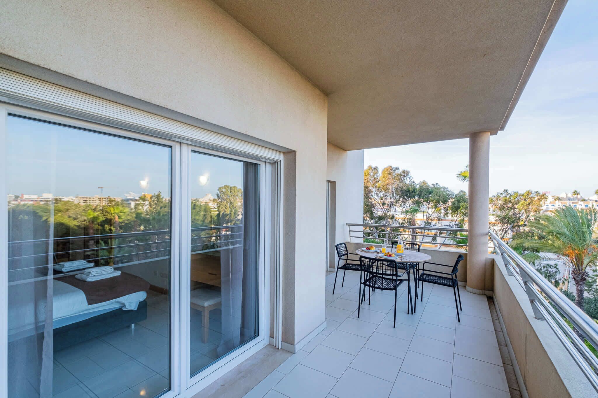 Ein Balkon mit Esstisch und Stühlen, ein Bett mit Blick durch eine Glastür sowie ein Blick auf die Stadtlandschaft.