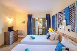 A hotel room with two beds, a wooden dresser, a television, a wall-mounted light, and a view of the outdoors through a sliding door.