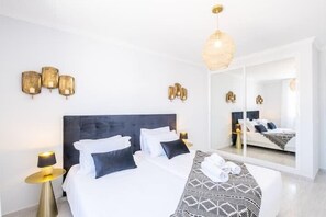 A modern hotel room with a large bed, a bedside table with a lamp, a mirror, and wall-mounted sconces.