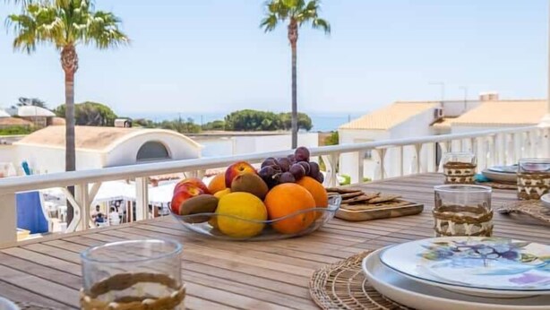 Un tavolo con una ciotola di frutta, bicchieri e un piatto su un pontile di legno, con vista su palme e edifici.